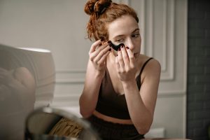 Red-haired woman applying an eye mask for skincare in a cozy room.
